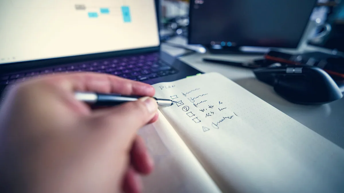 Une main qui écrit des notes à côté d'un ordinateur portable.